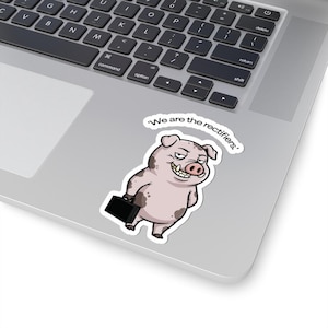 May include: A die-cut sticker featuring a cartoon pig with brown spots, a mischievous grin, and sharp teeth, holding a black briefcase. Above the pig, the text reads, 'We are the rectifiers.' The sticker is applied to the surface of a silver laptop.