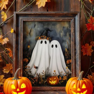 Puede incluir: Un cuadro enmarcado con temática de Halloween representa dos figuras de fantasmas blancos con ojos negros, uno con un sombrero de bruja. Dos calabazas iluminadas flanquean el cuadro, con hojas de otoño y un fondo de madera.