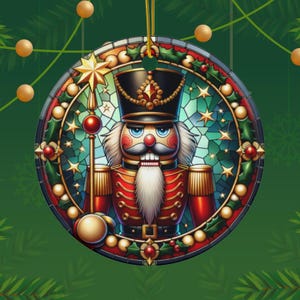 Puede incluir: Un adorno navideño redondo con un diseño de vitral colorido de un cascanueces. El cascanueces lleva un uniforme rojo y dorado con una cara azul y blanca. El adorno está rodeado de una corona de acebo verde y estrellas.
