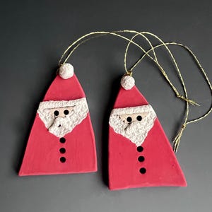 May include: Two red Santa Claus ornaments with white beards and hats, and black button details. Each ornament has a gold string for hanging. The ornaments are handmade and are perfect for Christmas decorations.