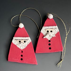 May include: Two red ceramic Christmas ornaments shaped like Santa Claus. Each ornament has a white beard and a white hat. The ornaments have gold string for hanging.