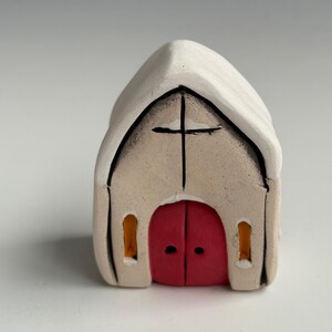 Könnte beinhalten: Kleine Keramik-Kirchenfigur mit weißem Dach und beigem Körper. Die Kirche hat eine rote Doppeltür und ein schwarzes Kreuz auf der Vorderseite. Zwei kleine, orange-gelbe Fenster flankieren die Tür.