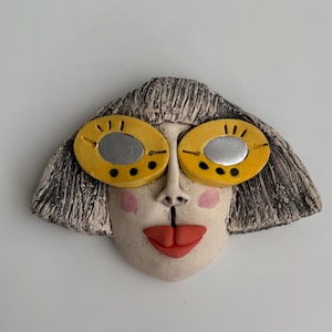 May include: A ceramic face sculpture with yellow sunglasses, red lips, and pink cheeks. The sculpture has a textured, gray hairstyle and a neutral skin tone. The sunglasses have silver centers and black dots.