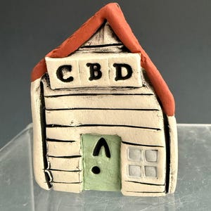 May include: A small, handmade ceramic house with a red roof and white walls. The house features a green door, a small window, and the letters "CBD" above the door. The house is a decorative item.