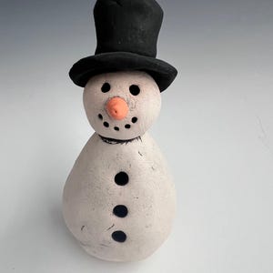 Könnte beinhalten: Eine kleine weiße Schneemann-Figur mit einem schwarzen Zylinder und einer orangefarbenen Karottennase. Der Schneemann hat drei schwarze Knöpfe.