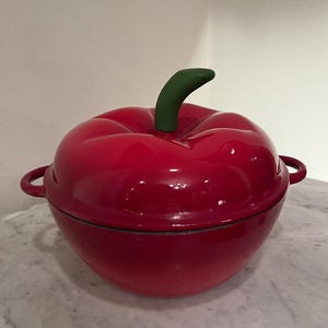 May include: A red, apple-shaped cast iron Dutch oven with a green stem handle. The pot has two small side handles and a glossy finish. The Dutch oven is sitting on a marble surface.