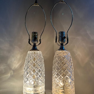 May include: Two vintage table lamps with clear, diamond-patterned glass bases and brass-colored metal accents. The lamps have a classic design with a bulbous shape and a metal frame for a lampshade. The lamps are lit, casting a warm glow.
