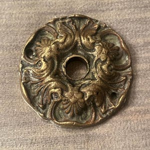 May include: A circular brass escutcheon plate featuring an ornate, raised relief design of symmetrical scrolling foliage and floral motifs. The metal has a weathered, darkened patina with a central circular opening and two small screw holes.