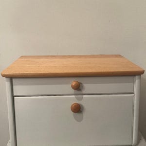 May include: A rectangular bread box featuring a light-colored wooden top and a matte white body. The front includes a small upper drawer and a larger lower drop-down door, each equipped with a single round, natural wood knob for easy opening.