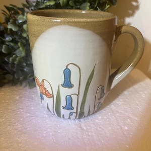 May include: A ceramic mug with a beige rim and handle, featuring a white body adorned with bluebell and butterfly illustrations. The mug has a light brown base and a curved handle, perfect for enjoying hot beverages.