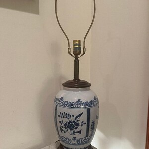 May include: A table lamp featuring a white ceramic base with blue floral patterns, a dark brown wooden base and top cap, and a brass harp fixture. The lamp body has a rounded, vase-like shape with intricate blue painted botanical designs.