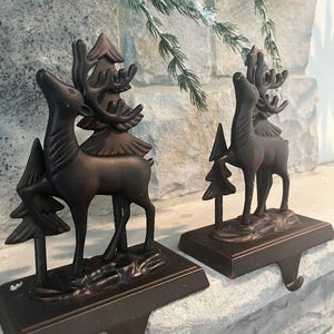 May include: A pair of dark bronze-toned metal stocking holders, each featuring a standing deer figure positioned in front of a stylized evergreen tree. The figures are mounted on rectangular bases with a hook extending from the front edge.
