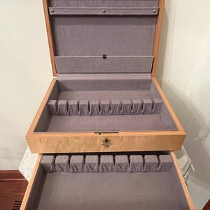 May include: A light-colored wooden chest with a hinged lid and a pull-out drawer. The interior is lined with gray fabric, featuring slotted organizers for flatware or utensils. A brass keyhole is centered on the front of the upper section.