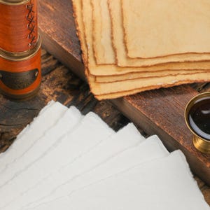 Op de afbeelding: Een stilleven in antieke stijl met stapels handgemaakt papier in wit en bruin, een kleine messing inktpot en een met leer omwikkeld spionnetje. De items zijn gerangschikt op een donker houten oppervlak.