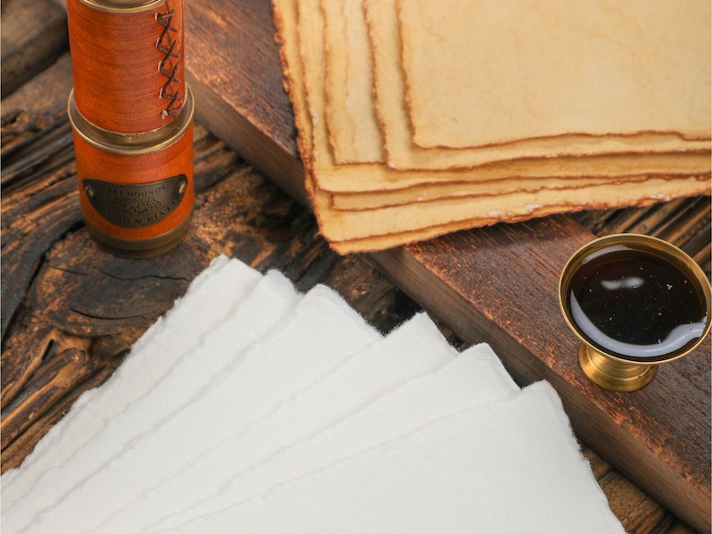 Op de afbeelding: Een stilleven in antieke stijl met stapels handgemaakt papier in wit en bruin, een kleine messing inktpot en een met leer omwikkeld spionnetje. De items zijn gerangschikt op een donker houten oppervlak.