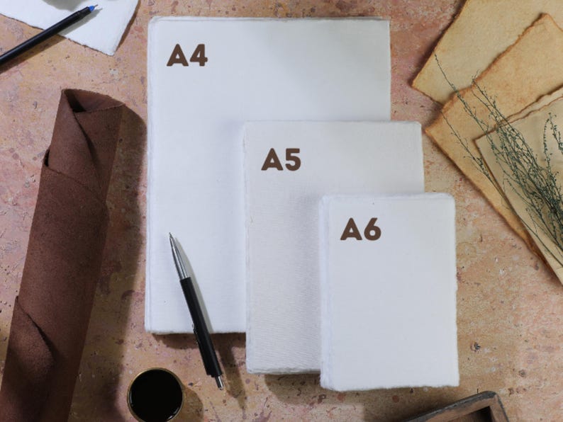 Op de afbeelding: Drie formaten wit papier, gelabeld als A4, A5 en A6, zijn op een getextureerd oppervlak gestapeld. Een pen, inkt en een opgerolde bruine papieren rol zijn ook aanwezig. Het papier lijkt handgemaakt te zijn, met ruwe randen.