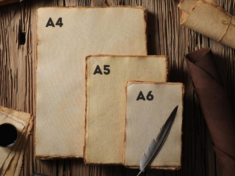 Op de afbeelding: Drie vellen handgemaakt papier in A4-, A5- en A6-formaat, met ruwe randen, zijn gerangschikt op een houten oppervlak. Een veerkiel en inktpot zijn ook aanwezig. Gerold papier en een gevouwen doek dragen bij aan de rustieke esthetiek.
