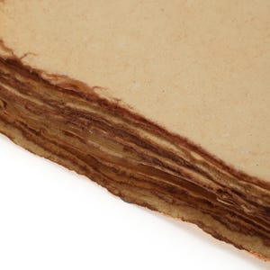 Op de afbeelding: Close-up van een stapel handgemaakt papier met ruwe, gescheurde randen. Het papier heeft een lichtbruine kleur op het oppervlak en een donkerdere bruine kleur op de randen, waardoor een gelaagd effect ontstaat. Het papier lijkt verouderd.