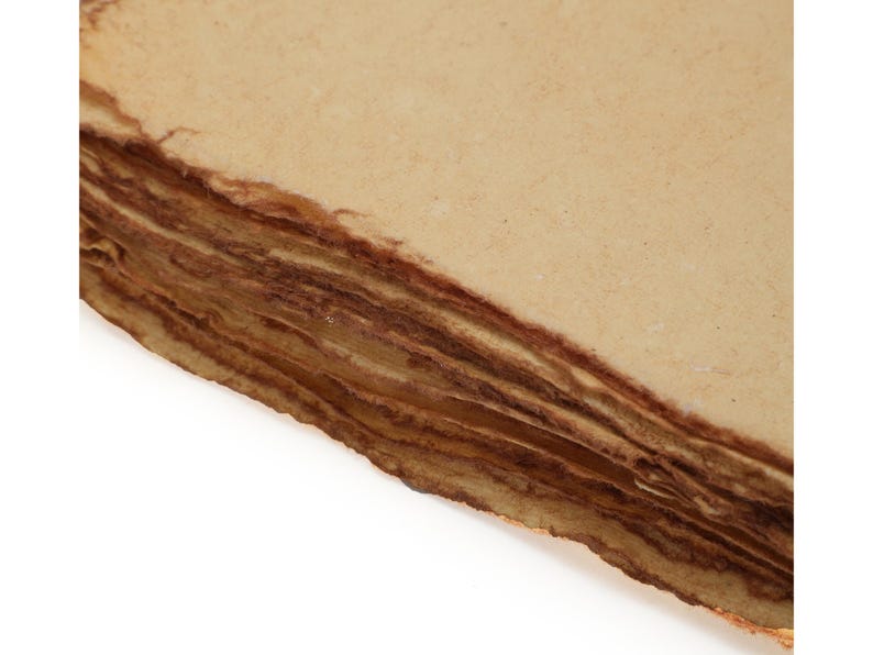 Op de afbeelding: Close-up van een stapel handgemaakt papier met ruwe, gescheurde randen. Het papier heeft een lichtbruine kleur op het oppervlak en een donkerdere bruine kleur op de randen, waardoor een gelaagd effect ontstaat. Het papier lijkt verouderd.