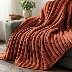 May include: A chunky knit throw blanket in a solid burnt orange color featuring a prominent vertical ribbed texture. The thick yarn creates deep, raised ridges throughout the fabric, which is draped over the arm of a gray upholstered sofa.
