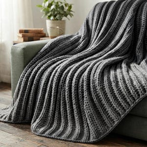 May include: A chunky knit throw blanket in a solid dark gray color featuring a prominent vertical ribbed texture. The blanket is draped over the arm and seat of a sofa, showcasing the thick yarn construction and consistent stitch pattern throughout.