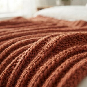 May include: A chunky knit throw blanket in a solid burnt orange color. The textile features a prominent vertical ribbed pattern created by thick yarn stitches, showcasing a soft, textured surface with deep folds and ridges throughout the fabric.