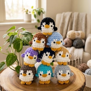May include: A stack of eight small, round, crocheted penguin plushies made from soft yarn. Each penguin features a white belly, black or colored wings and head, and small orange feet and beaks. The penguins are arranged in a pyramid formation.