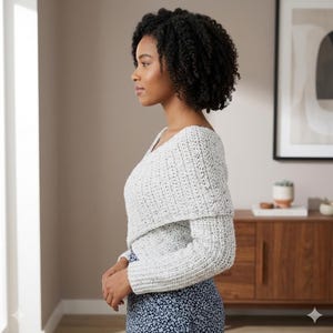 Könnte beinhalten: Ein hellgrauer, strukturierter Häkelpullover mit breitem, umgeschlagenem Kragen und langen Ärmeln. Der Pullover hat einen V-Ausschnitt und wird über einem blauen Rock mit Blumenmuster getragen. Das Model steht im Profil.