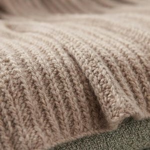 May include: A light beige knitted blanket featuring a prominent vertical ribbed texture. The fabric displays a consistent, repeating stitch pattern throughout, creating a soft, dimensional surface with deep, parallel ridges across the entire piece.