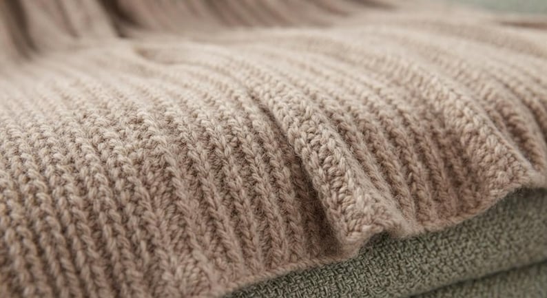 May include: A light beige knitted blanket featuring a prominent vertical ribbed texture. The fabric displays a consistent, repeating stitch pattern throughout, creating a soft, dimensional surface with deep, parallel ridges across the entire piece.