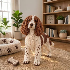 May include: A crocheted spaniel dog figurine with white and brown yarn, featuring long curly ears, a spotted coat, and a matching crocheted bone toy. The dog has dark eyes and a brown nose, standing on a textured surface near a pet bed.