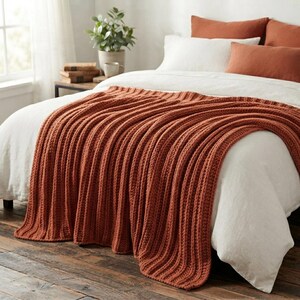 May include: A thick, rust-colored knitted throw blanket with a prominent vertical ribbed texture draped over a bed. The blanket features a chunky yarn construction and is layered over white bedding with matching rust-colored accent pillows.