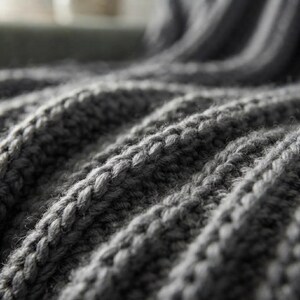 May include: A dark gray knitted blanket featuring a thick, textured ribbed pattern. The fabric displays a series of raised, vertical stitches that create deep folds and valleys across the surface, highlighting the chunky yarn construction.
