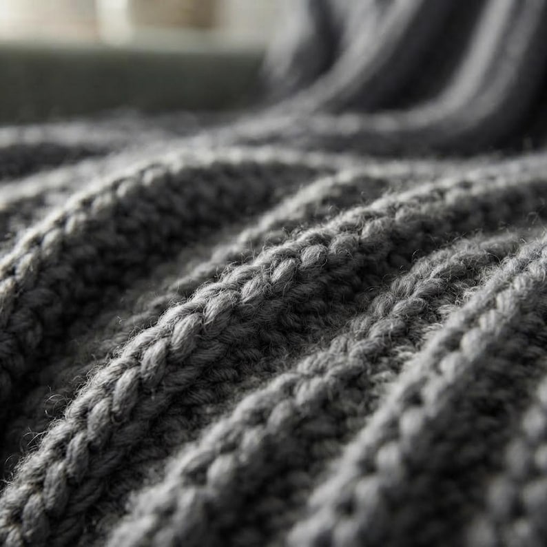 May include: A dark gray knitted blanket featuring a thick, textured ribbed pattern. The fabric displays a series of raised, vertical stitches that create deep folds and valleys across the surface, highlighting the chunky yarn construction.