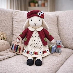 Può includere: Bambola coniglietto fatta a mano, lavorata a maglia, con giacca bordeaux, cappello e vestito a pois con dettagli floreali. La bambola tiene in mano una piccola borsa ed è accompagnata da un gatto lavorato a maglia, libri e occhiali. La bambola è seduta su un divano beige.
