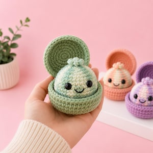 May include: A small, round, crocheted green creature with black bead eyes and a stitched smile, nestled inside a matching green crocheted clam-like shell container. The item features a textured yarn surface and a small decorative top knot.