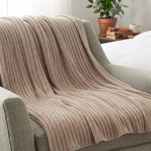 May include: A beige knitted throw blanket with a vertical ribbed texture draped over a light green upholstered armchair. The blanket features a consistent woven pattern throughout its length, providing a soft and textured appearance on the seat.