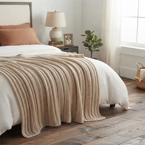 May include: A beige knitted throw blanket with a vertical ribbed texture draped over a white bedspread. The blanket features a consistent woven pattern and is positioned across the foot of the bed, contrasting with the white bedding and pillows.