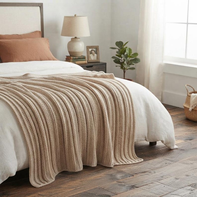 May include: A beige knitted throw blanket with a vertical ribbed texture draped over a white bedspread. The blanket features a consistent woven pattern and is positioned across the foot of the bed, contrasting with the white bedding and pillows.