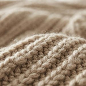 May include: A close-up view of a beige knitted fabric featuring a prominent ribbed texture. The knit pattern consists of vertical columns of raised stitches that create a soft, dimensional surface with a consistent light tan color throughout the textile.