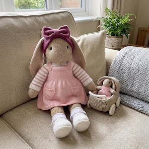 May include: A large beige knitted rabbit doll wearing a pink dress with heart-shaped pockets, a striped long-sleeve shirt, white socks, and a dark pink bow headband, accompanied by a smaller matching rabbit doll inside a beige knitted toy stroller.