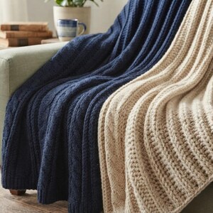 May include: A pair of thick, knitted blankets draped over a sofa. One blanket is a deep navy blue with a vertical cable knit texture, while the other is a light beige with a prominent ribbed knit pattern, both featuring a soft, chunky yarn construction.