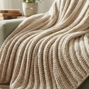 May include: A beige crochet throw blanket featuring a prominent vertical ribbed texture. The blanket is draped over a sage green sofa, showcasing thick yarn loops and a consistent knit pattern throughout the entire length of the fabric.