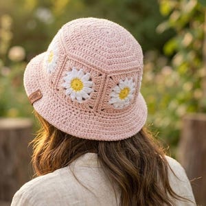 May include: A light pink crochet bucket hat with three white and yellow daisy appliqués. The hat has a small brim and a brown leather tag. The hat is a handmade item.