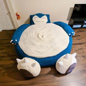 May include: A large, handmade blue and white crocheted Snorlax-shaped floor cushion with two matching footrests. The cushion features a smiling face on the headrest and a cream-colored belly. The footrests are cream, brown, and white.