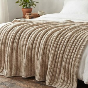 May include: A beige knitted throw blanket with a prominent vertical ribbed texture draped over a white bedspread. The blanket features a thick, chunky yarn construction with consistent parallel lines running the length of the fabric.