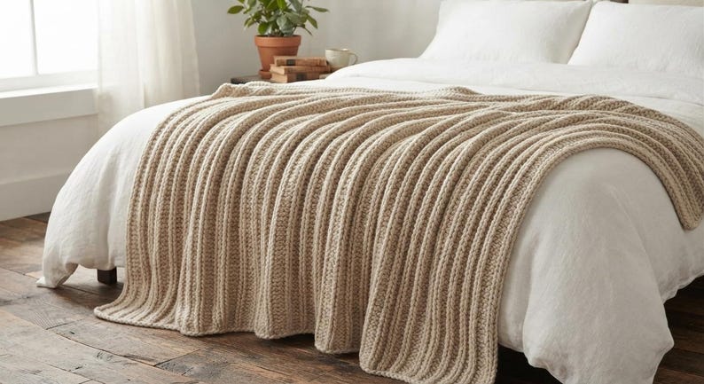 May include: A beige knitted throw blanket with a prominent vertical ribbed texture draped over a white bedspread. The blanket features a thick, chunky yarn construction with consistent parallel lines running the length of the fabric.