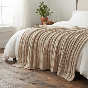 May include: A beige knitted throw blanket with a prominent vertical ribbed texture draped over a white bed. The blanket features a thick, chunky yarn construction with consistent parallel lines running the length of the fabric, creating a soft, layered appearance.