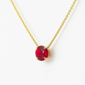 Op de afbeelding: Een goudkleurige ketting met een levendige rode, ovaal geslepen edelsteen hanger. De hanger is gezet in een gouden bezel en hangt aan een fijne gouden ketting. De ketting wordt getoond tegen een witte achtergrond.
