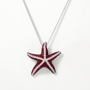 Puede incluir: Un collar de plata con un colgante de estrella de mar. La estrella de mar es roja con detalles blancos y negros. El colgante está hecho de pequeñas piedras preciosas. El collar está sobre un fondo blanco.